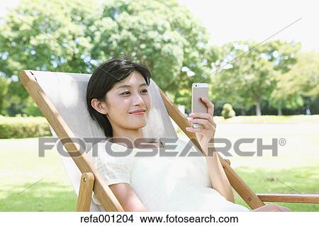Japanese woman with smartphone on deck chair in a city park View Large Photo Image Picture - Japanese woman with smartphone on deck chair in a city park. Fotosearch