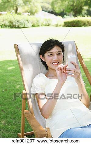 Japanese woman with smartphone on deck chair in a city park View Large Photo Image Stock Photo - Japanese woman with smartphone on deck chair in a city park. Fotosearch
