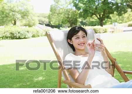 Japanese woman with smartphone on deck chair in a city park View Large Photo Image Stock Photograph - Japanese woman with smartphone on deck chair in a city park. Fotosearch
