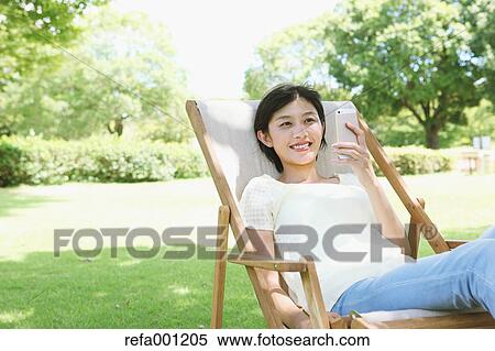 Stock Photography - Japanese woman with smartphone on deck chair in a city park. Fotosearch