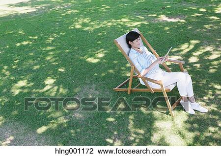 Stock Image - Japanese woman with tablet on deck chair in a city park. Fotosearch
