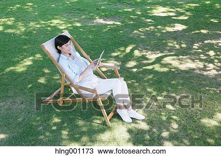Stock Image - Japanese woman with tablet on deck chair in a city park. Fotosearch