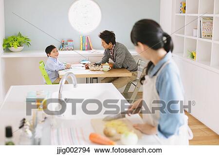 Mother cooking in an open kitchen watching father and son doing homework at the dining table View Large Photo Image Picture - Mother cooking in an open kitchen watching father and son doing homework at the dining table. Fotosearch