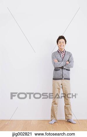 Stock Image - Young adult man standing in front of a white wall with crossed arms. Fotosearch