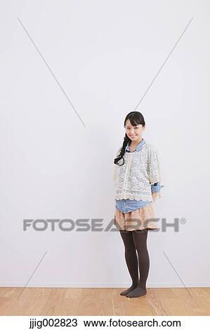Stock Image - Young adult woman leaning against a white wall and smiling at camera. Fotosearch