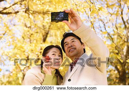 Stock Image - Senior Japanese couple taking pictures in a city park in Autumn. Fotosearch