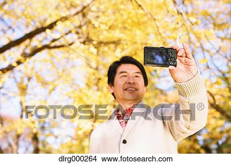 Picture - Senior Japanese man taking a selfie in a city park in Autumn. Fotosearch