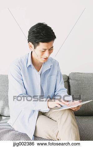 Stock Photograph - Young Japanese man with tablet on the sofa. Fotosearch