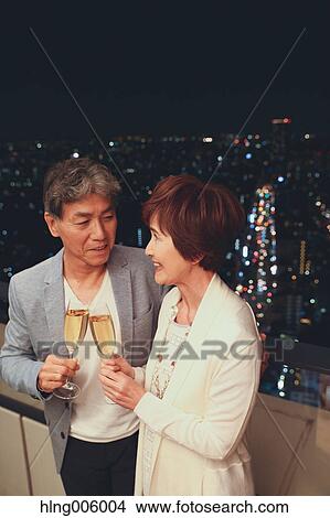 Picture - Fashionable Japanese senior couple toasting in front of night cityscape. Fotosearch