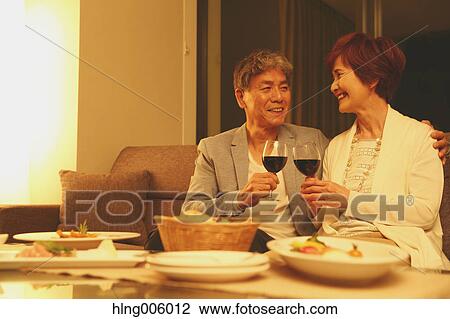 Stock Image - Fashionable Japanese senior couple toasting on sofa. Fotosearch