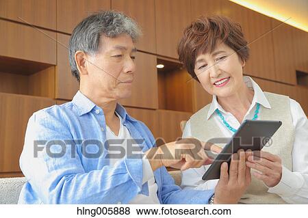 Stock Photo - Fashionable Japanese senior couple with tablet on sofa. Fotosearch