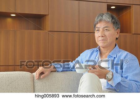 Stock Image - Fashionable Japanese senior man having a cup of coffee on the sofa. Fotosearch