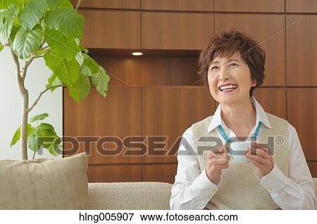 Stock Photo - Fashionable Japanese senior woman having a cup of coffee on the sofa. Fotosearch