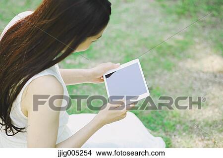 Picture - Portrait of young Japanese woman laying on grass with tablet. Fotosearch