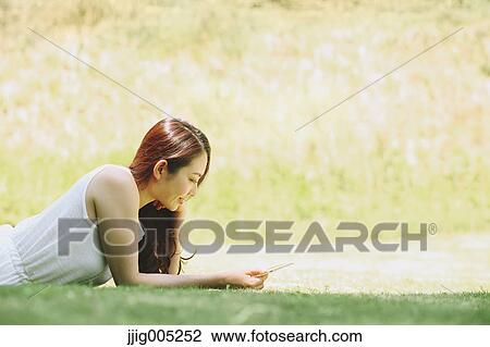 Stock Image - Portrait of young Japanese woman laying on grass with tablet. Fotosearch