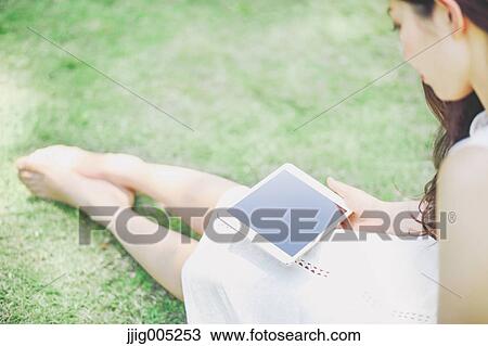 Stock Image - Portrait of young Japanese woman laying on grass with tablet. Fotosearch