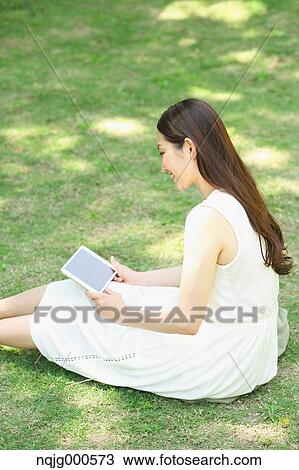 Stock Image - Portrait of young Japanese woman laying on grass with tablet. Fotosearch