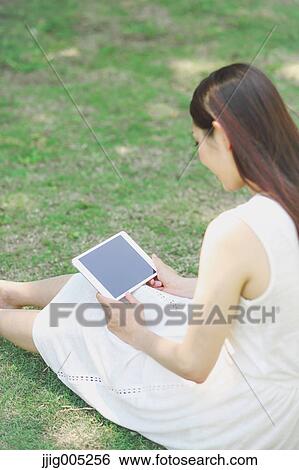 Stock Photograph - Portrait of young Japanese woman laying on grass with tablet. Fotosearch