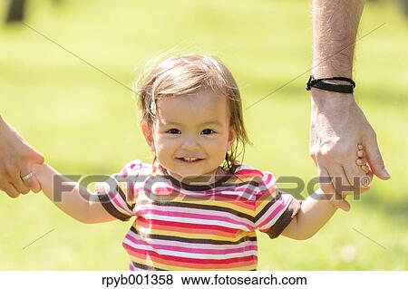 Toddler holding parents hands in a park View Large Photo Image Stock Photo - Toddler holding parents hands in a park. Fotosearch