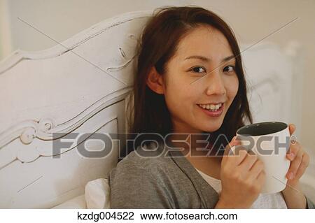 Stock Image - Young attractive Japanese woman drinking coffee in bed. Fotosearch