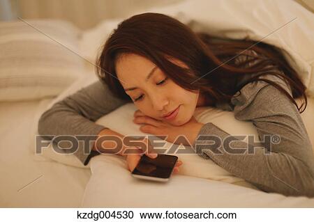 Stock Image - Young attractive Japanese woman with smartphone in bed. Fotosearch