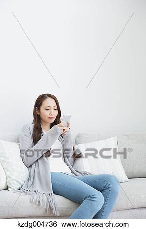 Stock Photograph - Young attractive Japanese woman with smartphone on the sofa. Fotosearch
