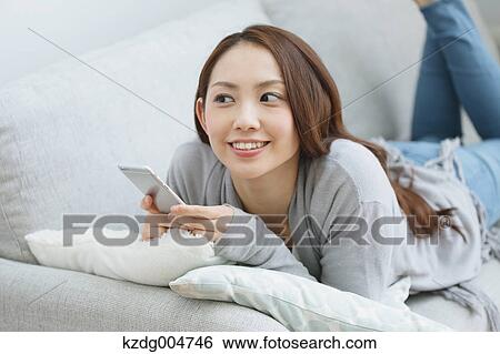 Stock Photograph - Young attractive Japanese woman with smartphone on the sofa. Fotosearch