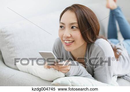 Stock Photography - Young attractive Japanese woman with smartphone on the sofa. Fotosearch
