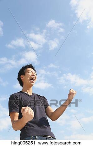 Stock Image - Young Japanese man cheering against blue sky. Fotosearch
