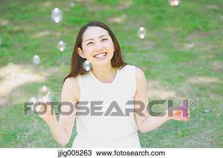 Stock Image - Young Japanese woman blowing soap bubbles in a city park. Fotosearch