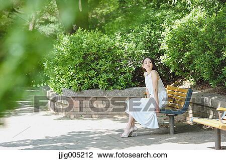 Stock Image - Young Japanese woman sitting on a bench in a city park. Fotosearch