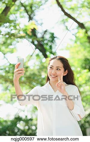 Stock Photo - Young Japanese woman taking selfie surrounded by green in a city park. Fotosearch