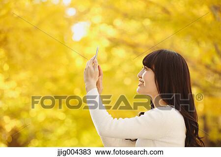 Stock Image - Young Japanese woman with smartphone in a city park. Fotosearch