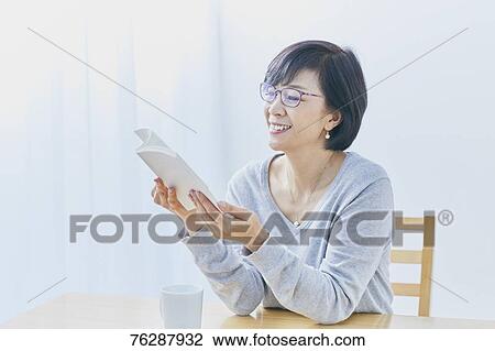 Stock Image - Japanese senior woman reading book at table. Fotosearch