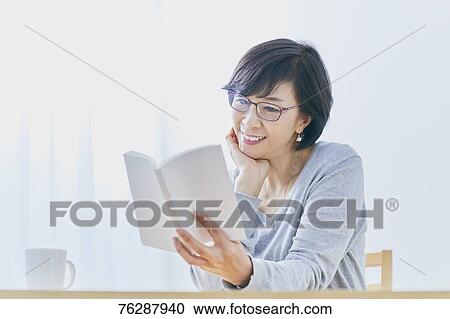 Stock Image - Japanese senior woman reading book at table. Fotosearch