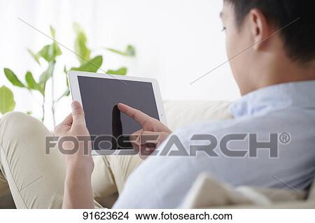 Picture - Young Japanese man using tablet on the sofa. Fotosearch