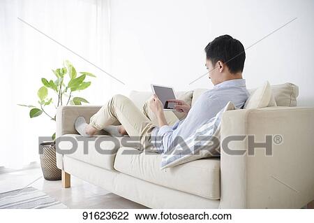 Stock Image - Young Japanese man using tablet on the sofa. Fotosearch
