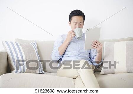 Stock Photo - Young Japanese man using tablet on the sofa. Fotosearch