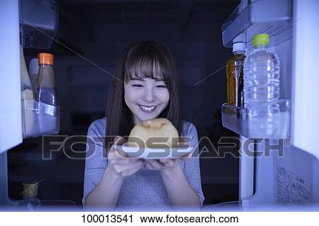 Stock Image - Young Japanese woman eating night snack from the fridge. Fotosearch