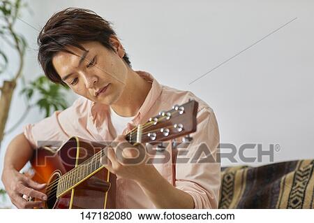 Stock Image - Japanese man playing guitar at home. Fotosearch