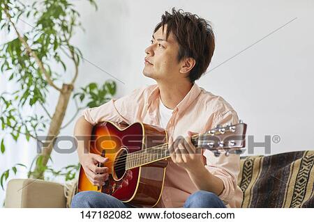 Stock Image - Japanese man playing guitar at home. Fotosearch