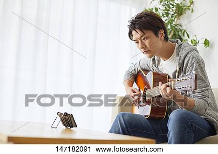 Stock Image - Japanese man playing guitar at home. Fotosearch