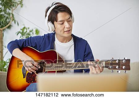 Stock Image - Japanese man playing guitar at home. Fotosearch