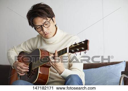 Stock Image - Japanese man playing guitar at home. Fotosearch