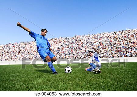 Stock Image - Soccer player kicking ball as defender slides in. Fotosearch