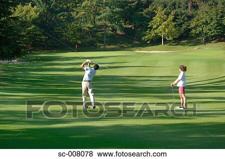 Male golfer hitting down the fairway as a woman looks on View Large Photo Image Stock Photo - Male golfer hitting down the fairway as a woman looks on. Fotosearch