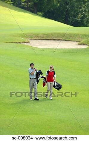 Picture - Side view of a couple walking on the fairway as they carry their golf bags. Fotosearch