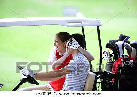Stock Image - A woman covers the eyes of her partner as he drives a golf cart. Fotosearch