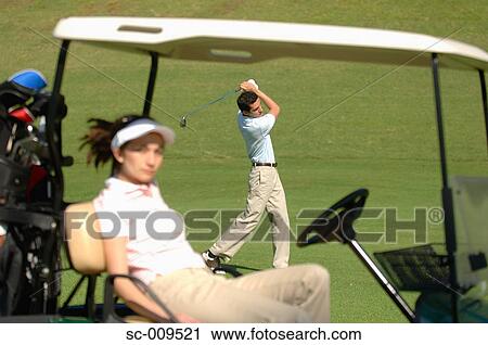 Stock Image - Female golfer catching some sunshine whilst her partner plays a shot. Fotosearch