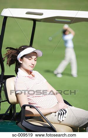 Stock Photo - Female golfer catching some sunshine whilst her partner plays a shot. Fotosearch